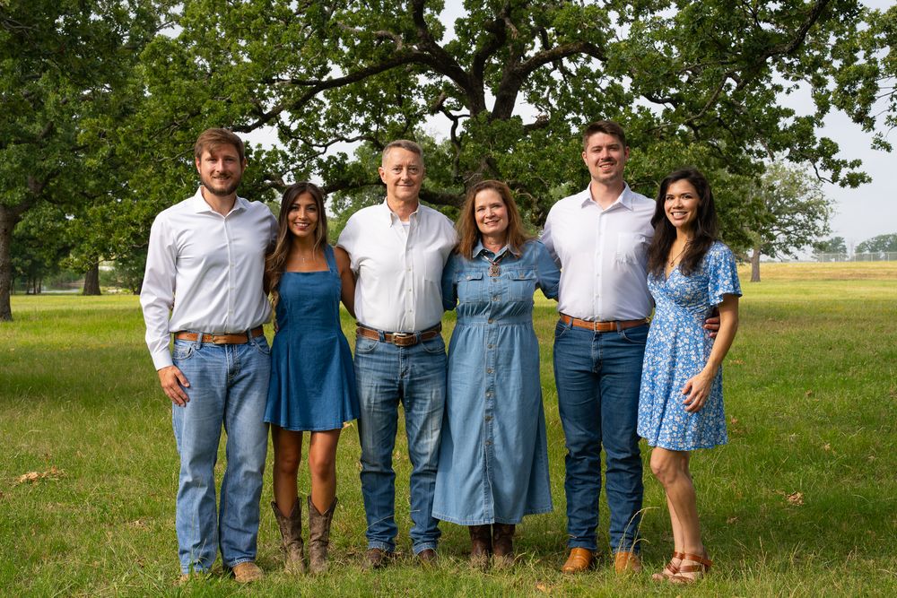 A group of six people stand together outdoors wearing casual summer attire in front of trees and a grassy field.