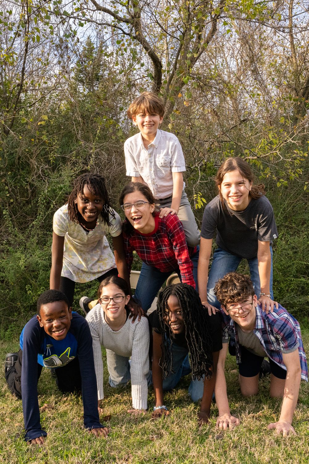 A diverse group of students create a playful human pyramid formation outdoors on grass with trees in the background.