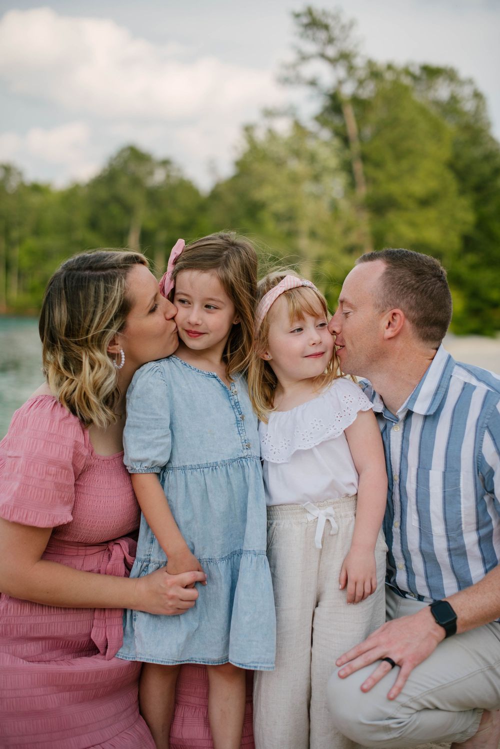 Family shares sweet kisses and cuddles during an outdoor photo session by the water on a summer day.