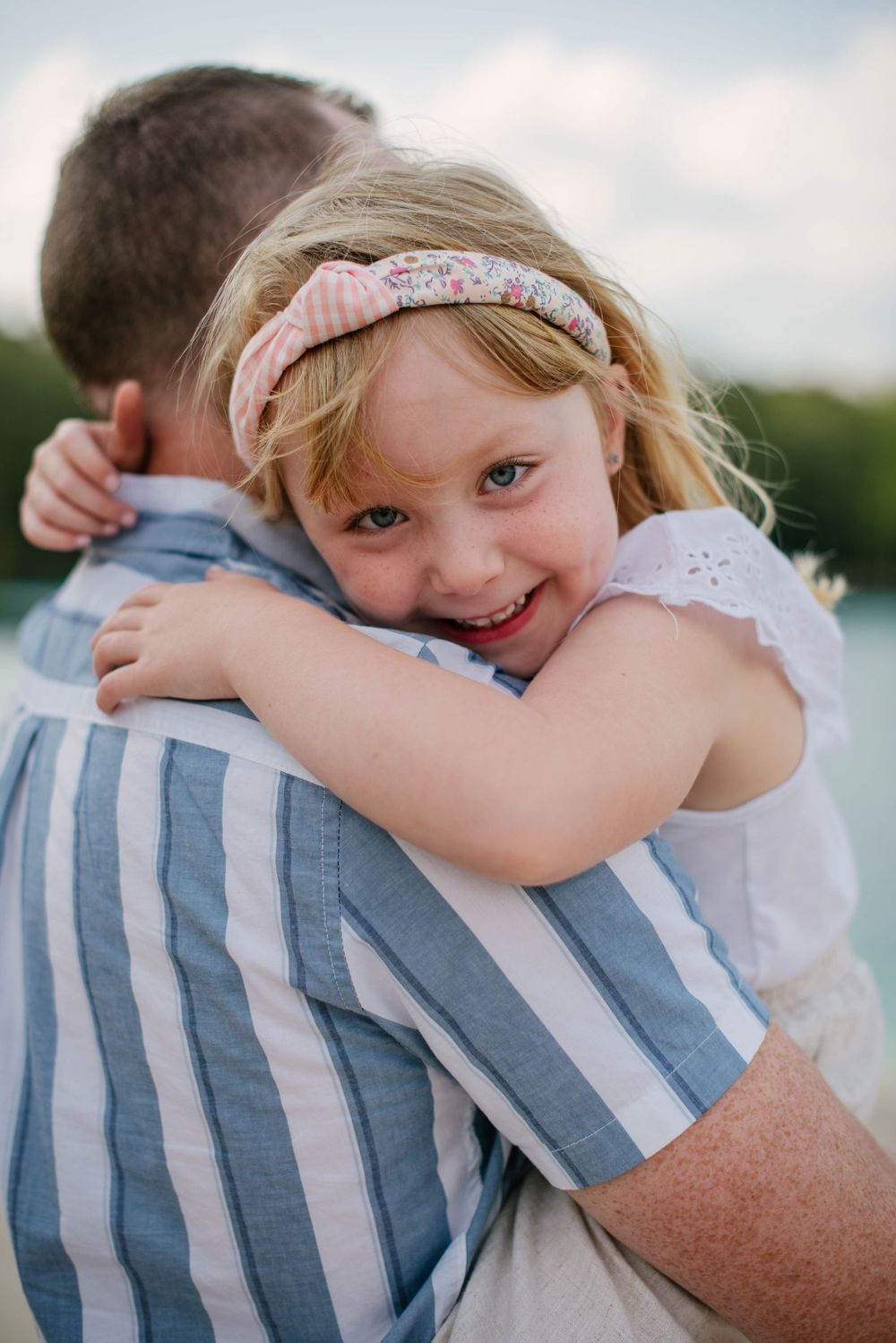 A small child in pink dress and floral headband hugs someone wearing blue and white striped clothing outdoors.