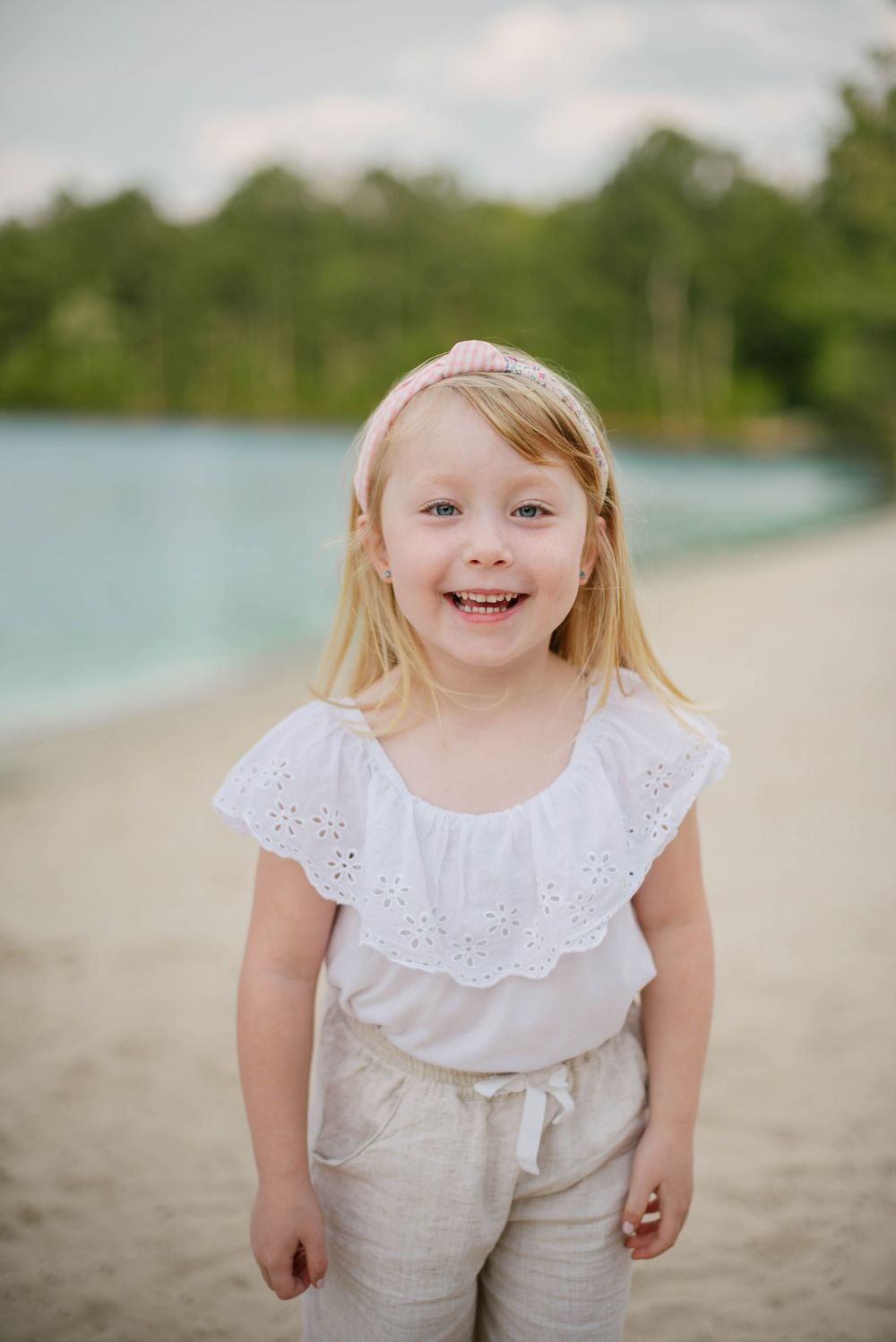Child wearing a white eyelet top and linen pants stands smiling on a beach with water and trees in the background.