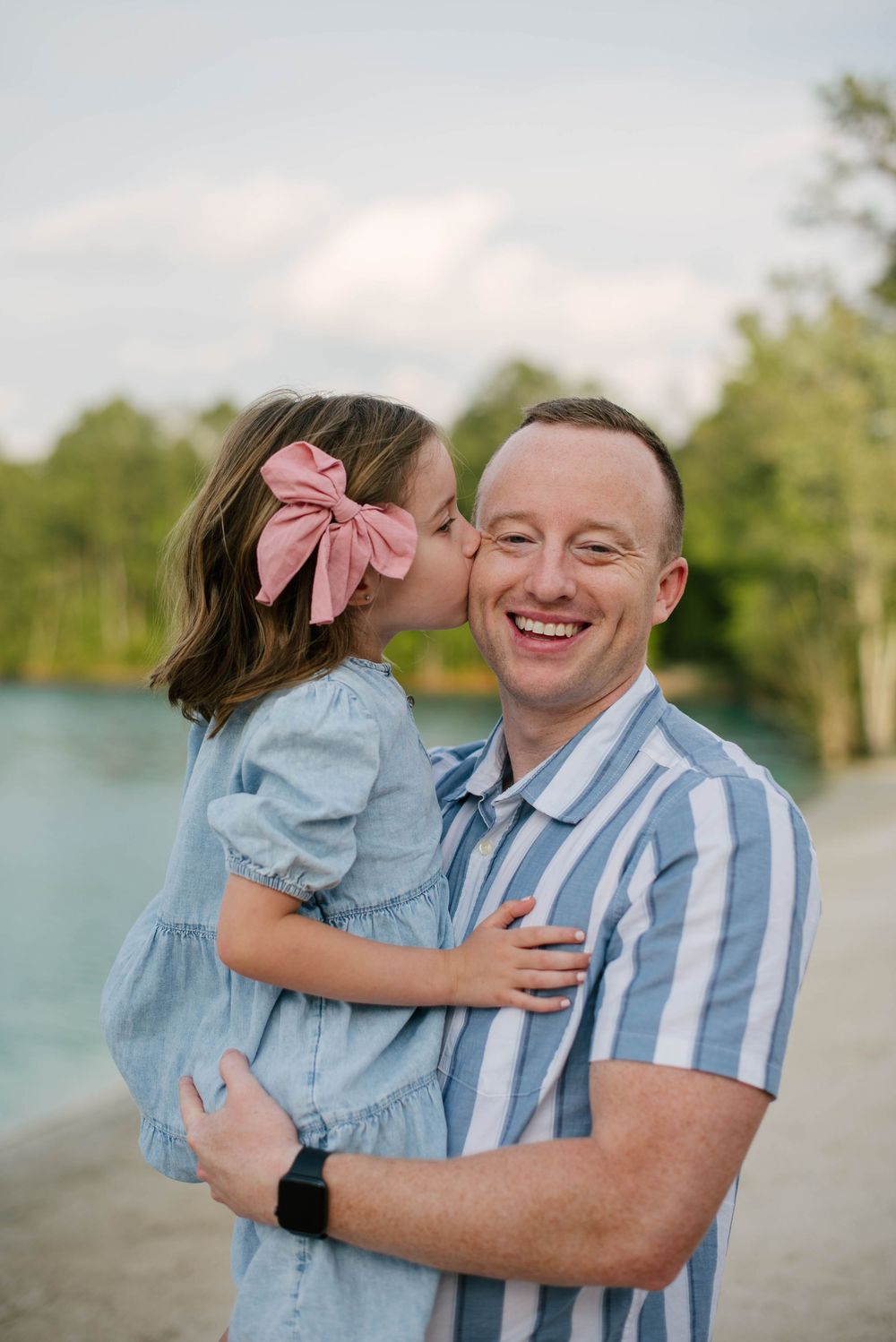 A parent and young child in matching blue outfits share an affectionate moment by a lake on a sunny day.