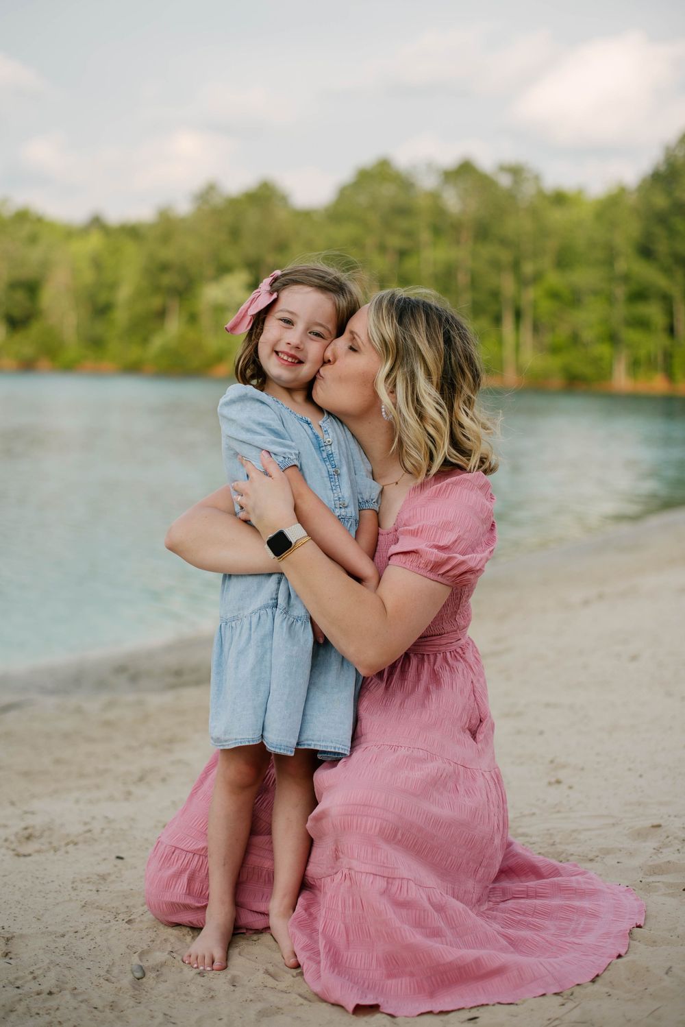 A tender moment shared between parent and child on a sandy beach with a serene lake and trees in the background.