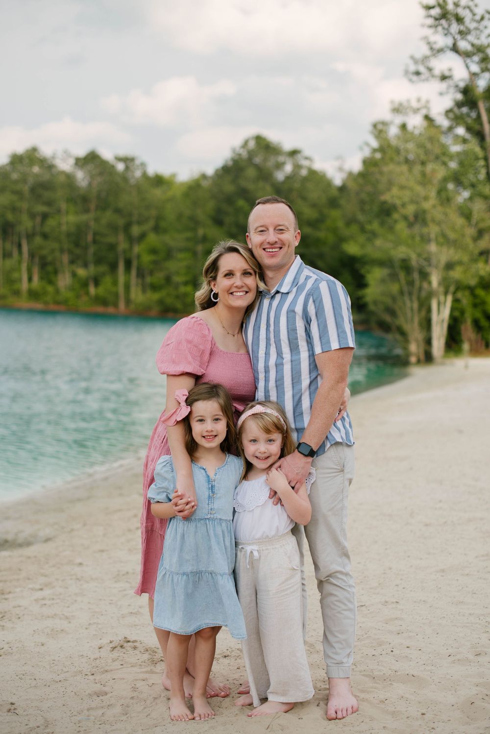 A family of four poses together on a sandy beach with turquoise water and green trees in the background.