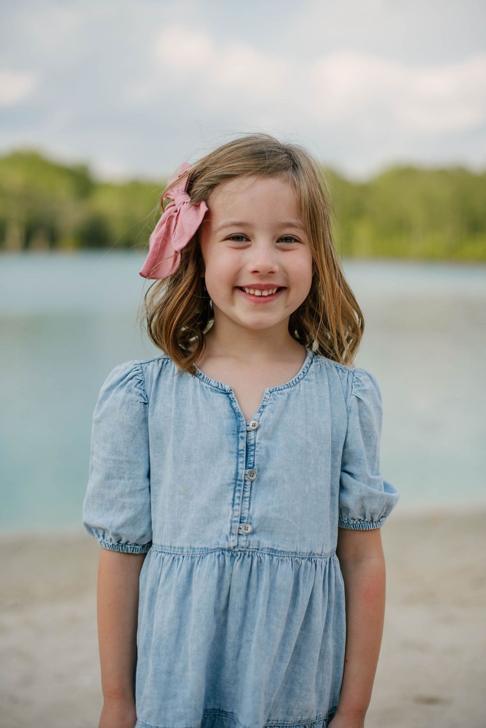 A young child in a chambray dress poses on a beach with water and trees in the background.