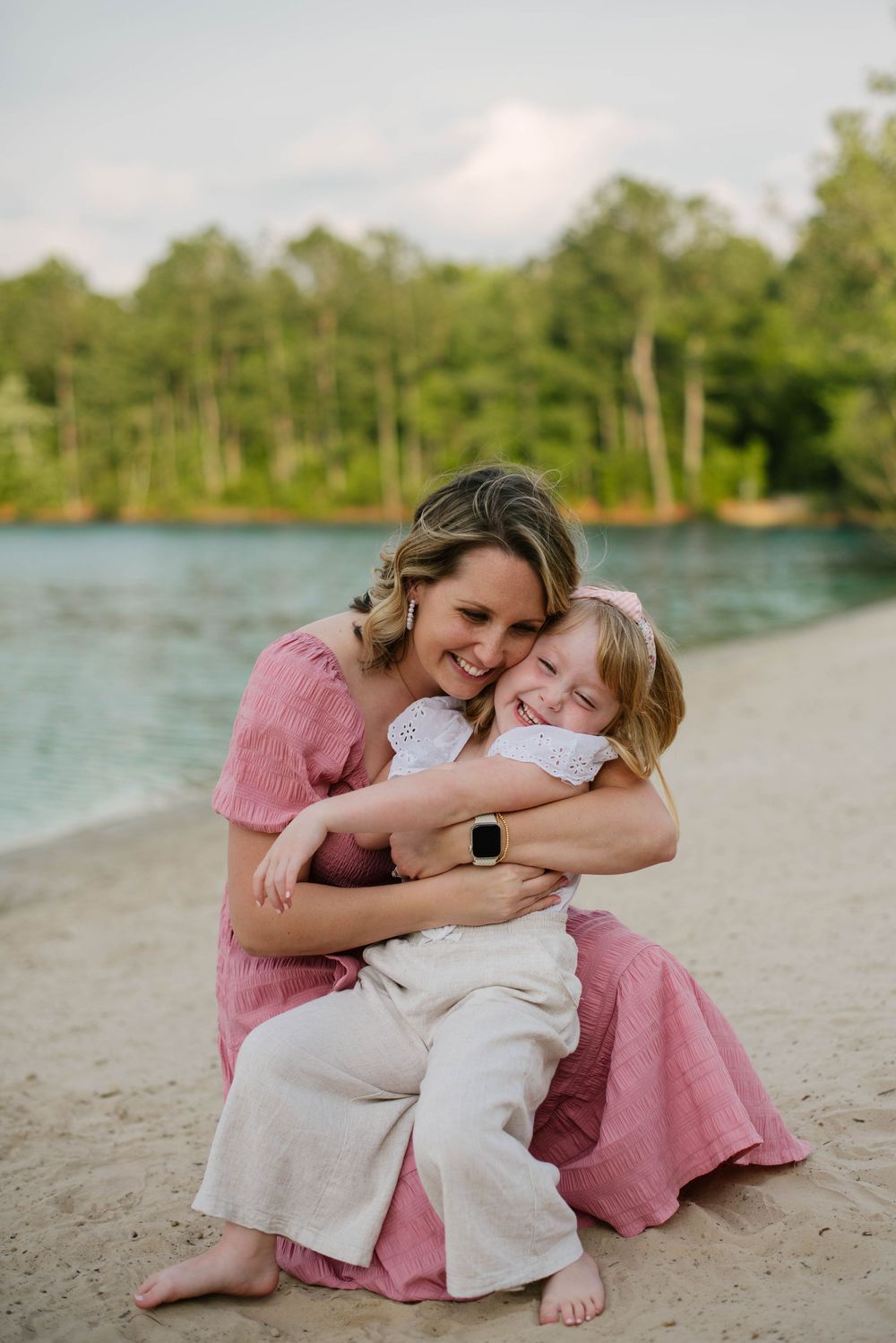 A tender moment shared between two people embracing on a sandy beach with a lake and trees in the background.