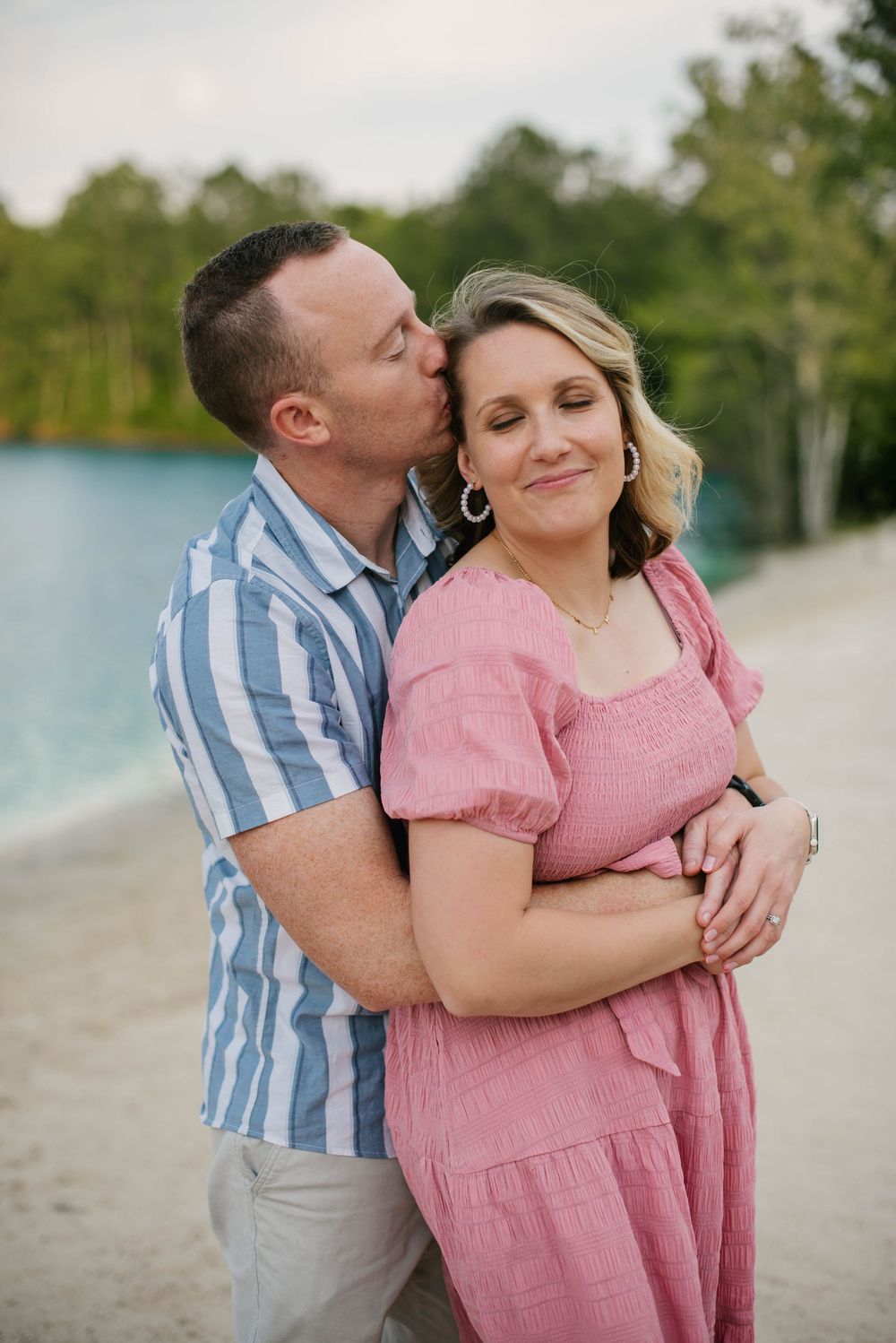 A romantic couple embrace on a beach path, with her in a pink dress and him in a blue striped shirt.