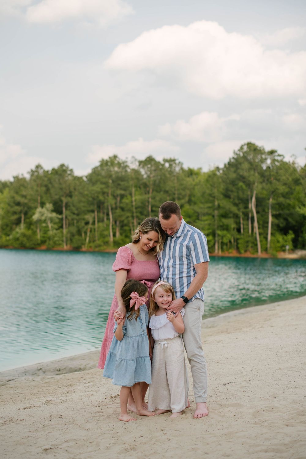 A family shares a tender moment together on a sandy beach with turquoise water and green trees in the background.