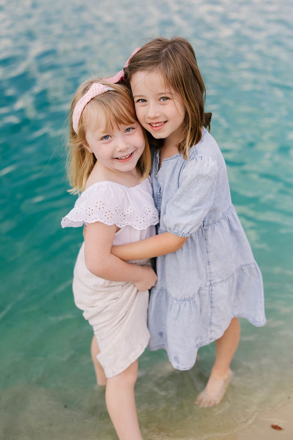 Two young girls in summer dresses hug and smile while playing at the edge of turquoise waters on a beach.
