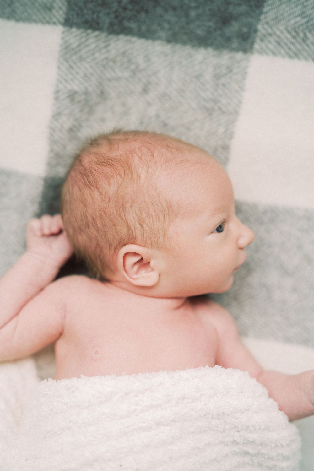 A newborn baby rests on a plaid blanket in profile view.