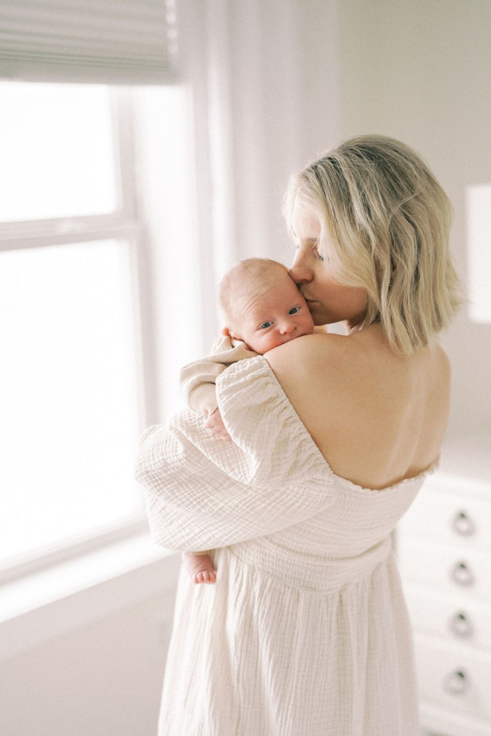Tender moment by window with parent in white dress holding newborn.