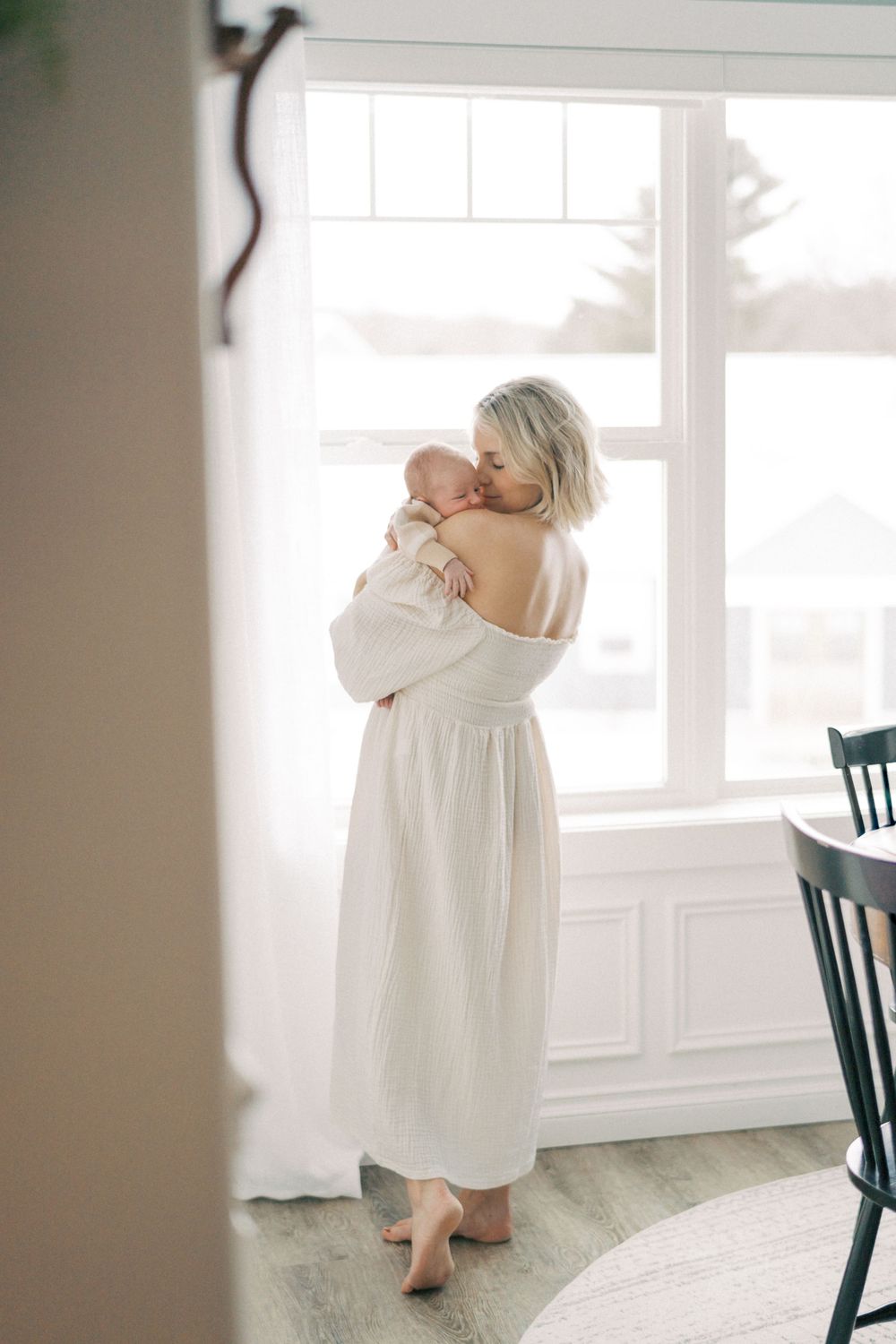Serene portrait by window showing parent in white dress holding baby.