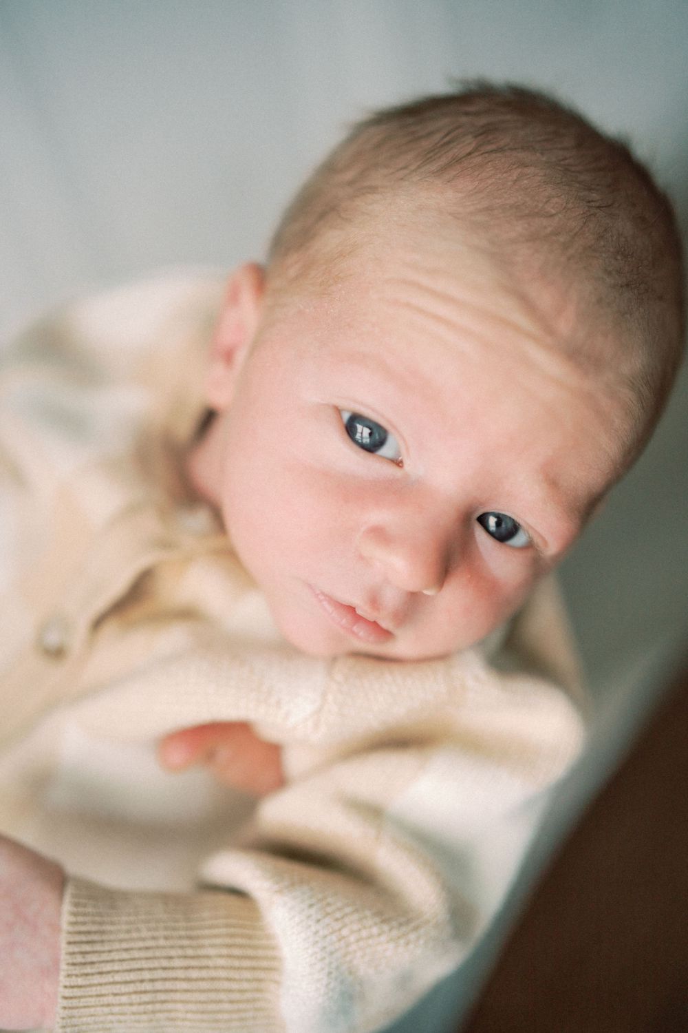 Close-up portrait of a newborn baby with bright eyes wearing a cream sweater against a soft background.