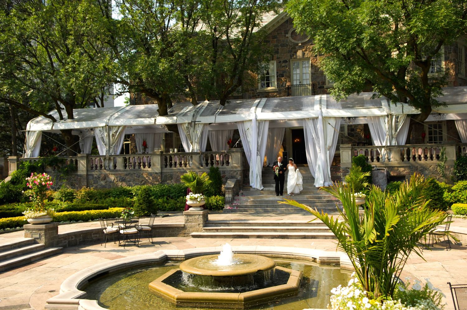 An elegant outdoor wedding venue with white drapes, fountain and tropical landscaping.