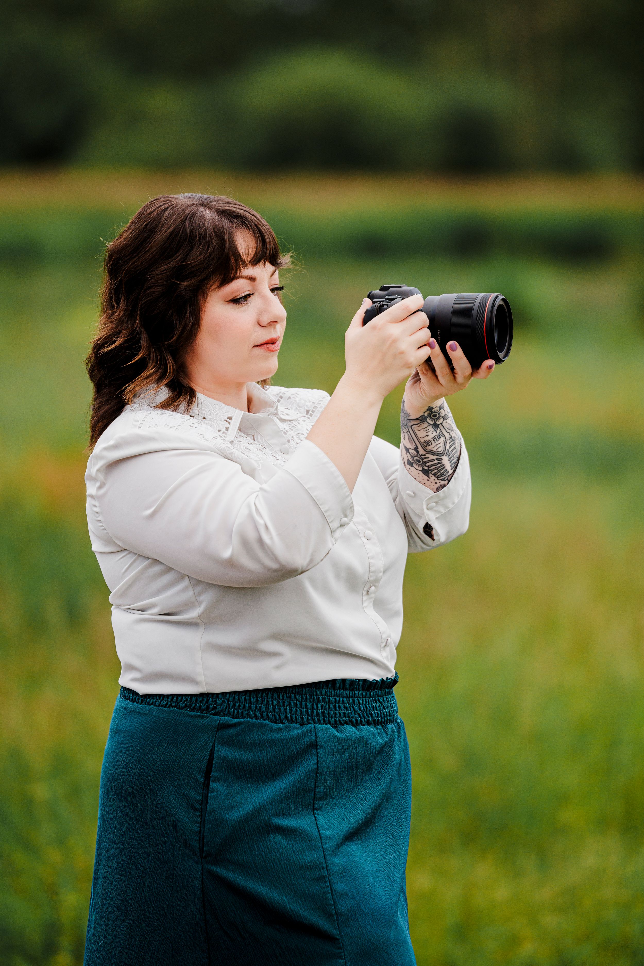 Photographer in a black floral sweater and distressed jeans sits barefoot holding a camera against an orange background.