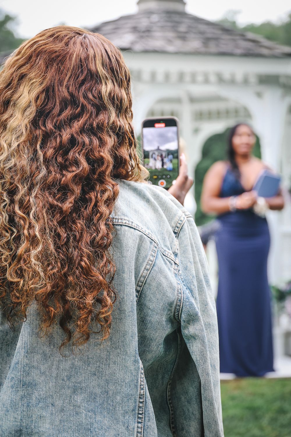 Someone with long curly brown hair wearing denim jacket takes photo of person in blue dress near white gazebo.