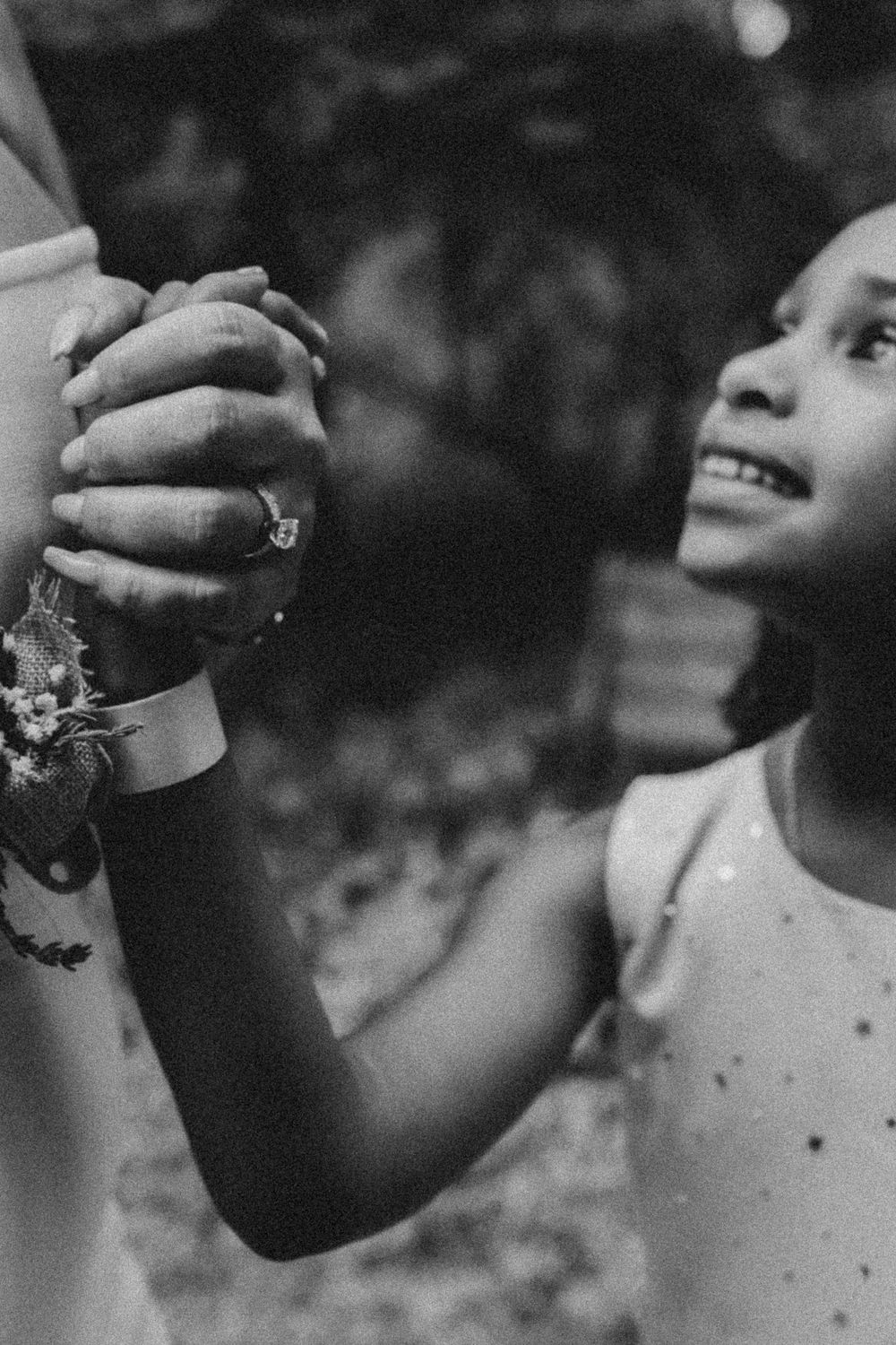 Black and white close-up of clasped hands with a corsage and a joyful smile in soft focus.