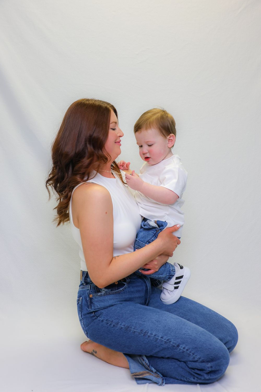 A parent in blue jeans plays and interacts lovingly with a baby wearing white against a light background.