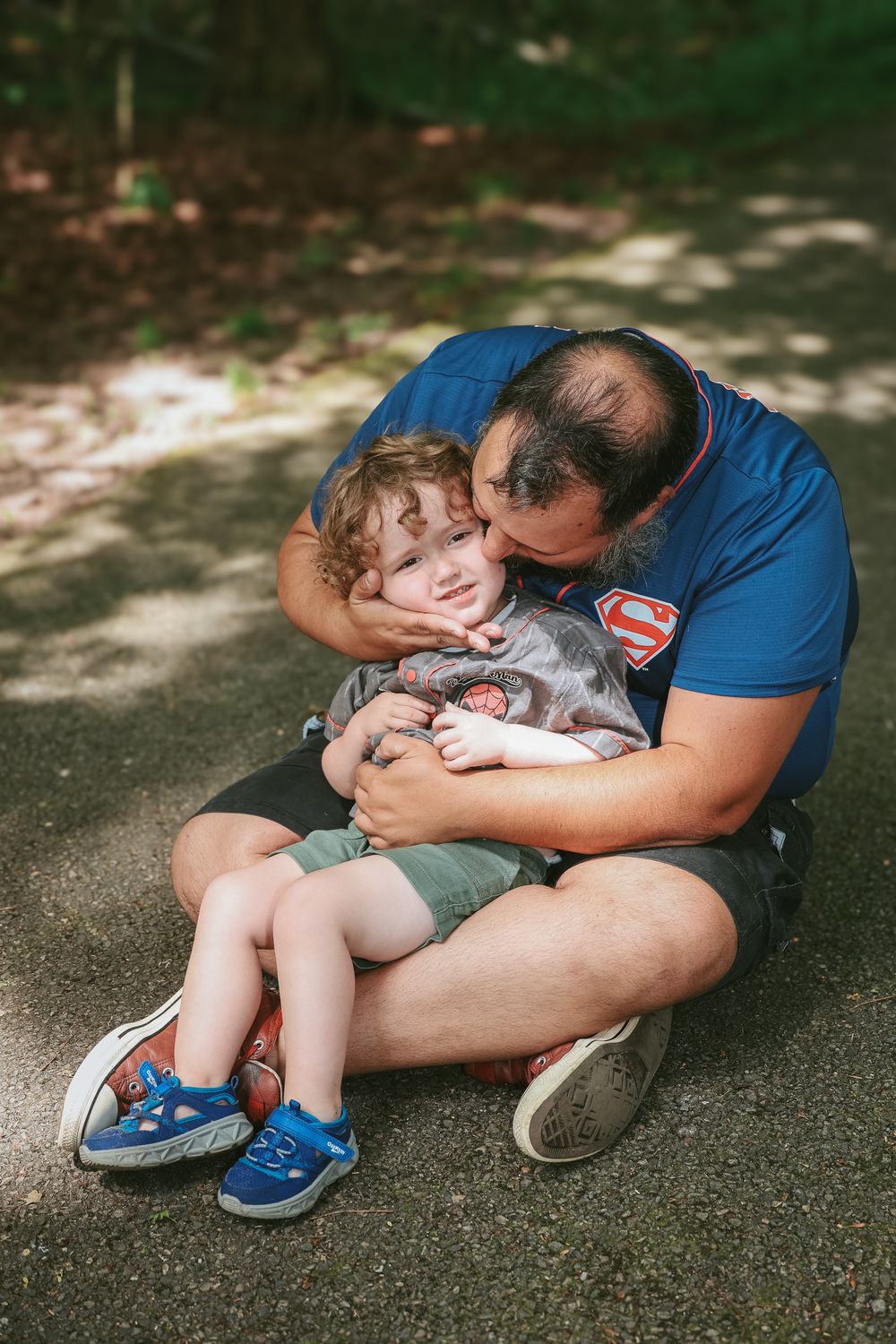 A parent in a Superman shirt sits on the ground hugging and playing with a young child on a sun-dappled path.