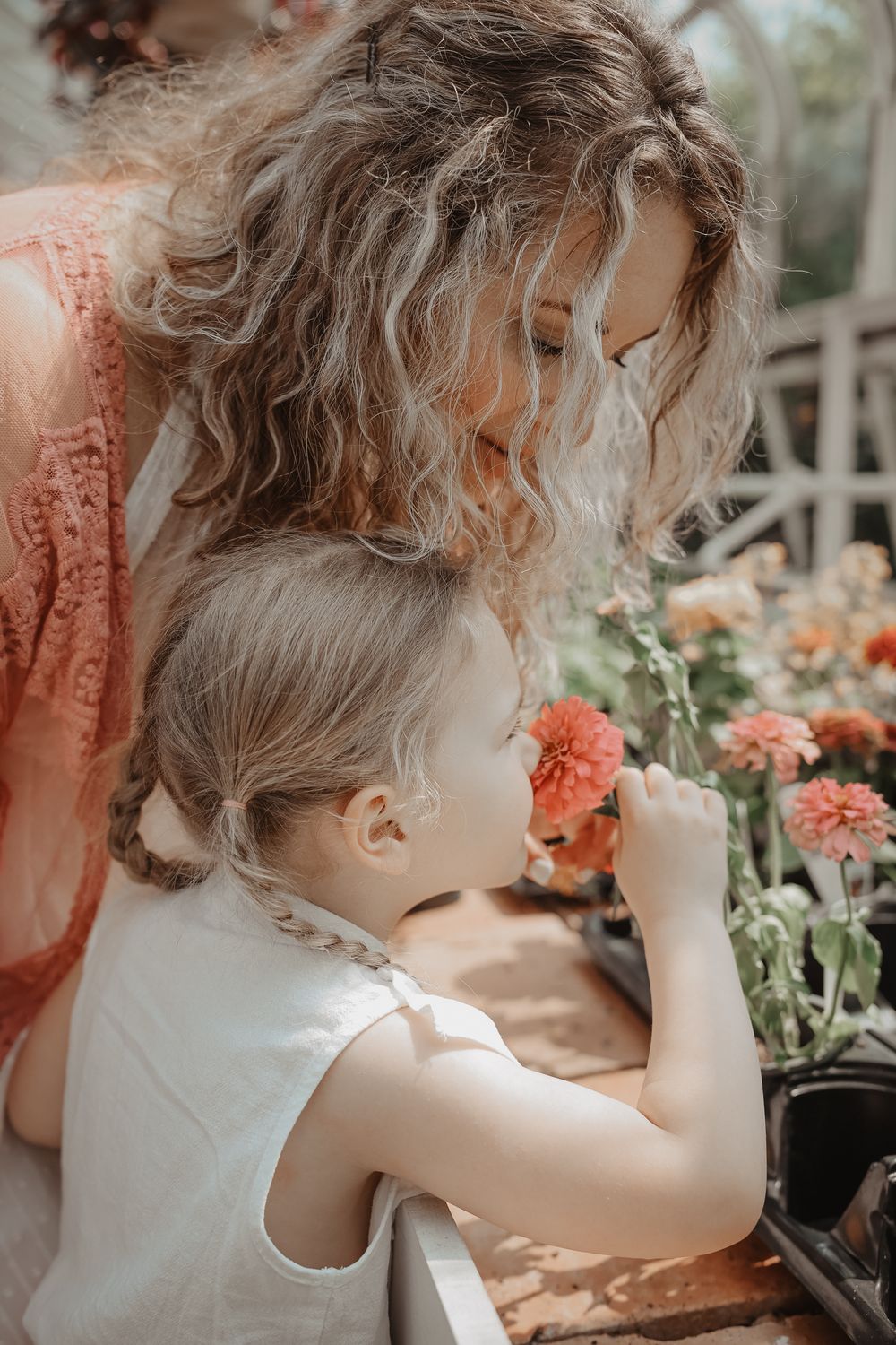 Curly-haired toddler in white dress explores blooming coral-colored flowers in a garden setting on a sunny day.