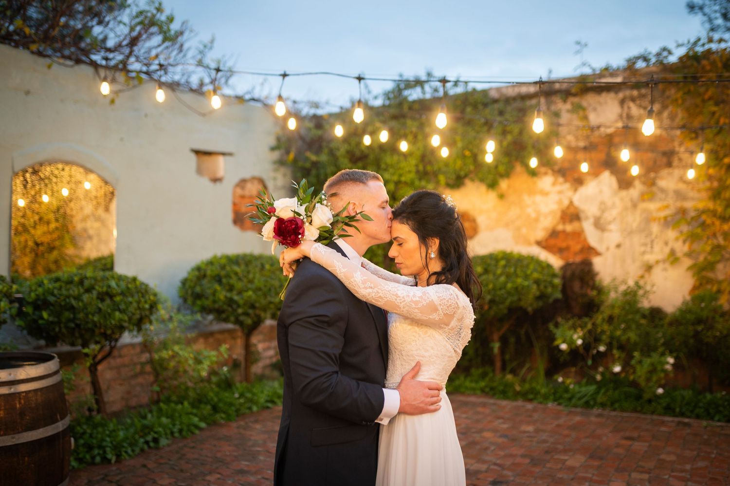 A couple dances beneath twinkling string lights in a romantic garden setting during sunset.
