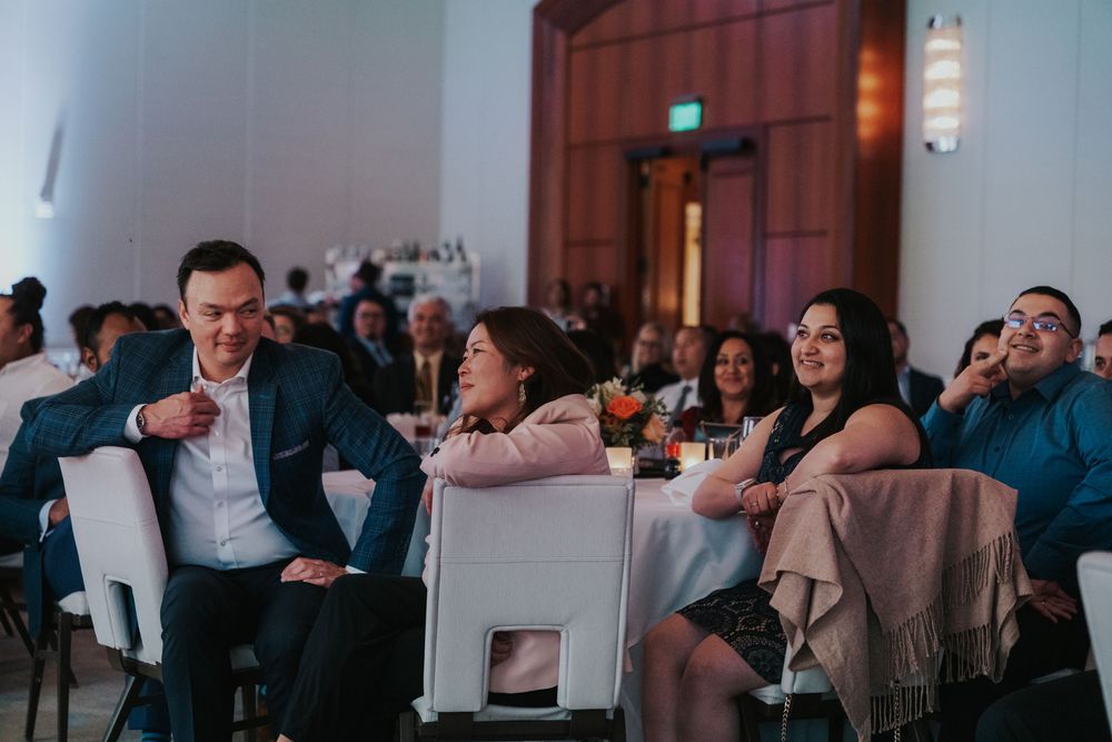 Wedding guests sit at decorated round tables during a reception, sharing conversation and laughter in a dimly lit venue.