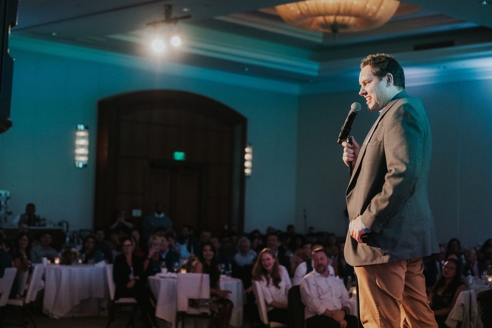 A speaker on stage performs at a dimly lit venue with blue lighting and round tables in the audience area.