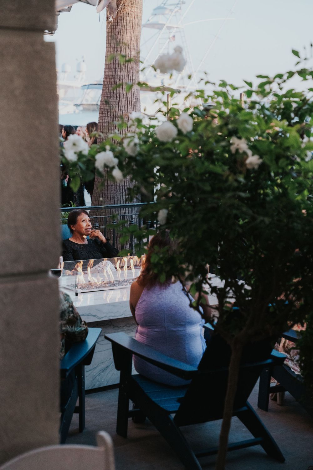 People sit at outdoor picnic tables surrounded by greenery at a restaurant patio on a summer evening.