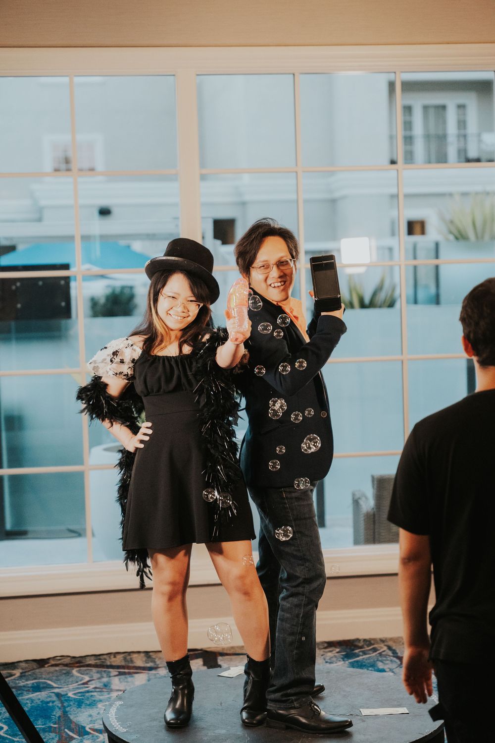Two people in black outfits pose together at an indoor event space with large windows and blue lighting.