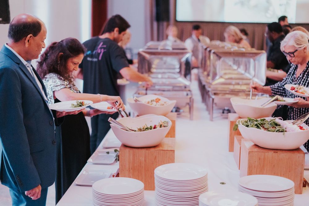 People serving themselves food from a buffet line with white plates and serving bowls on wooden tables.
