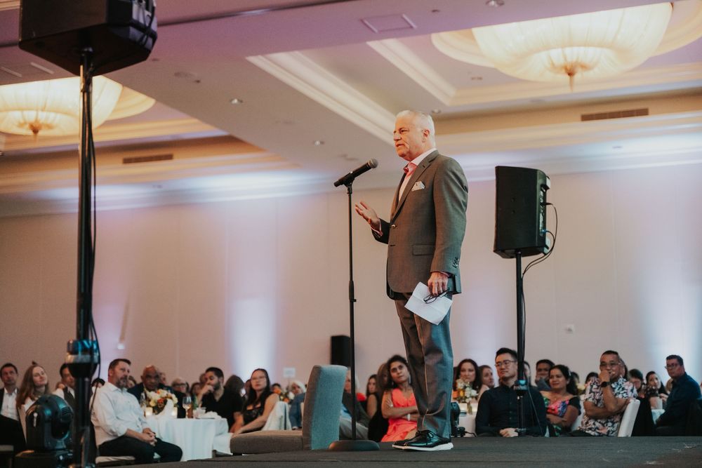 Speaker presents on stage at formal event with audience seated in conference room with ornate ceiling details.