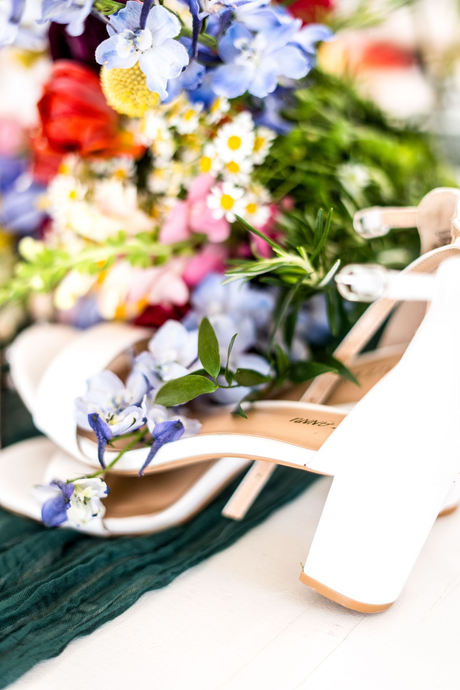 White wedding heels surrounded by delicate blue and colorful spring flowers on a green fabric backdrop.