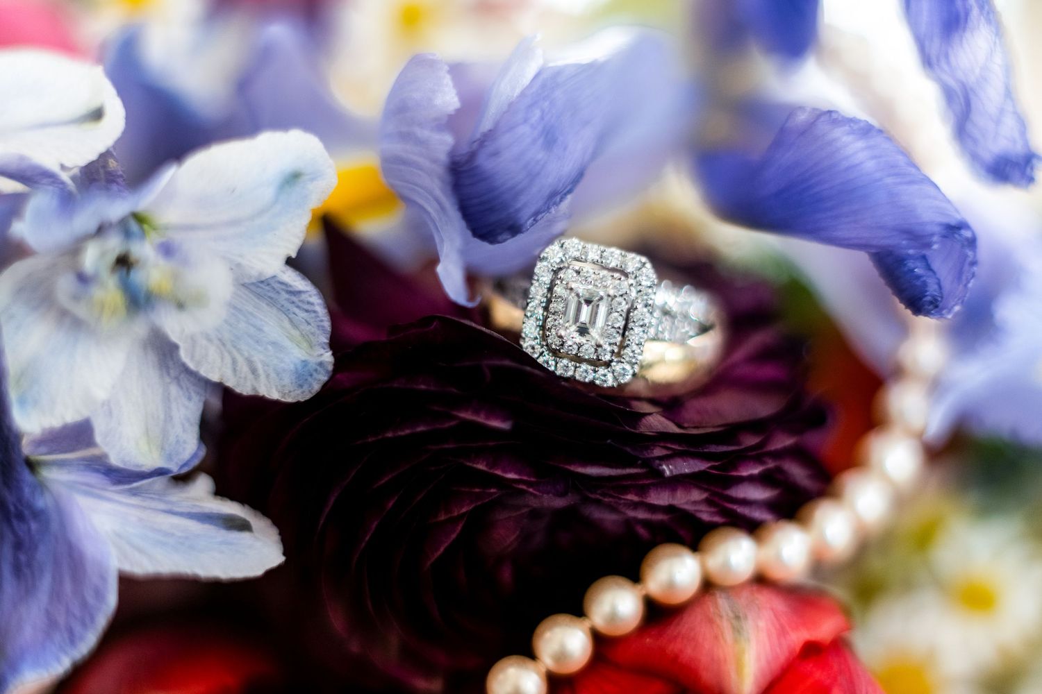 Close up view of an engagement ring with pearls and purple flowers on dark burgundy fabric.
