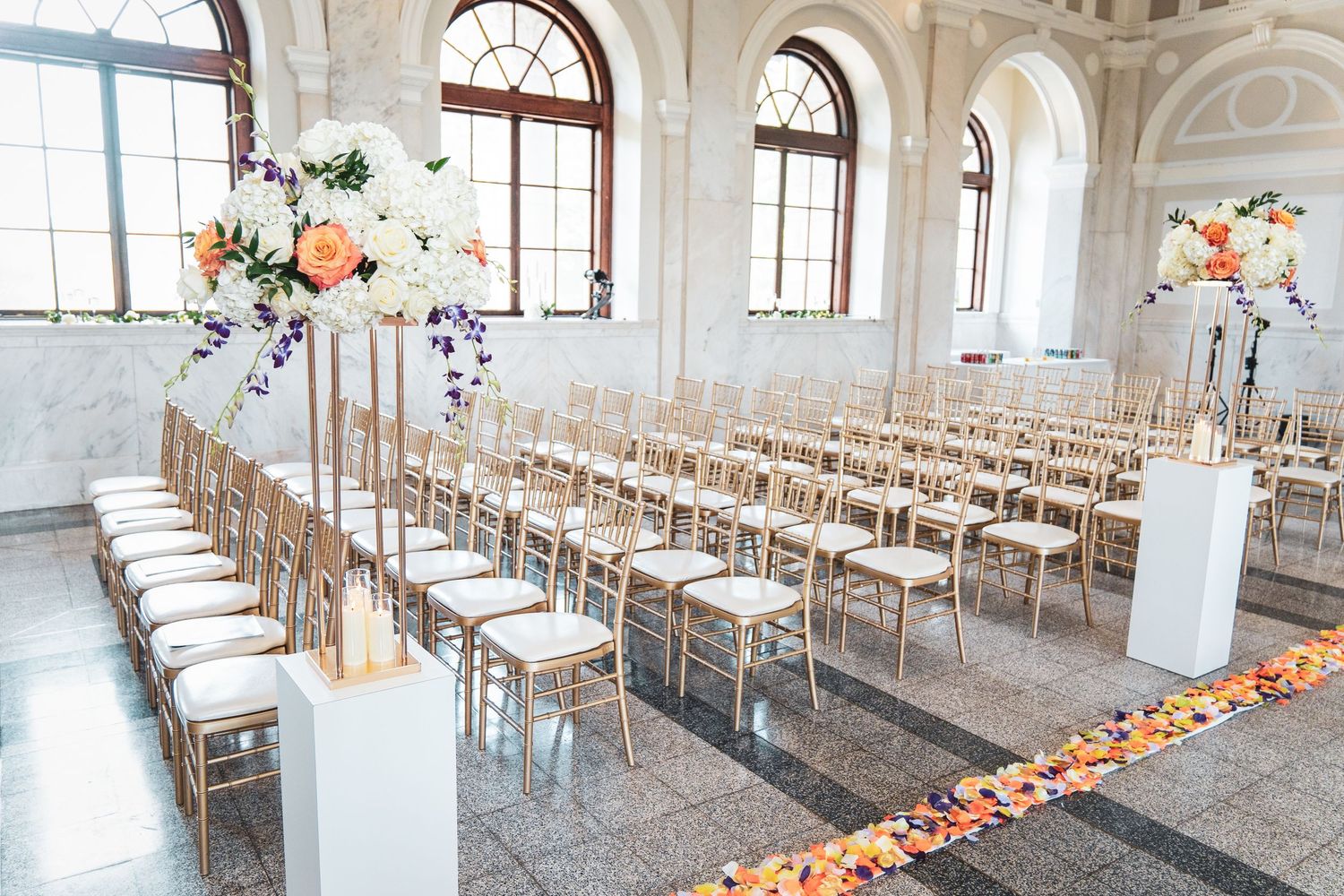 Elegant white ceremony space with gold chairs and tall floral arrangements in a bright room with arched windows.