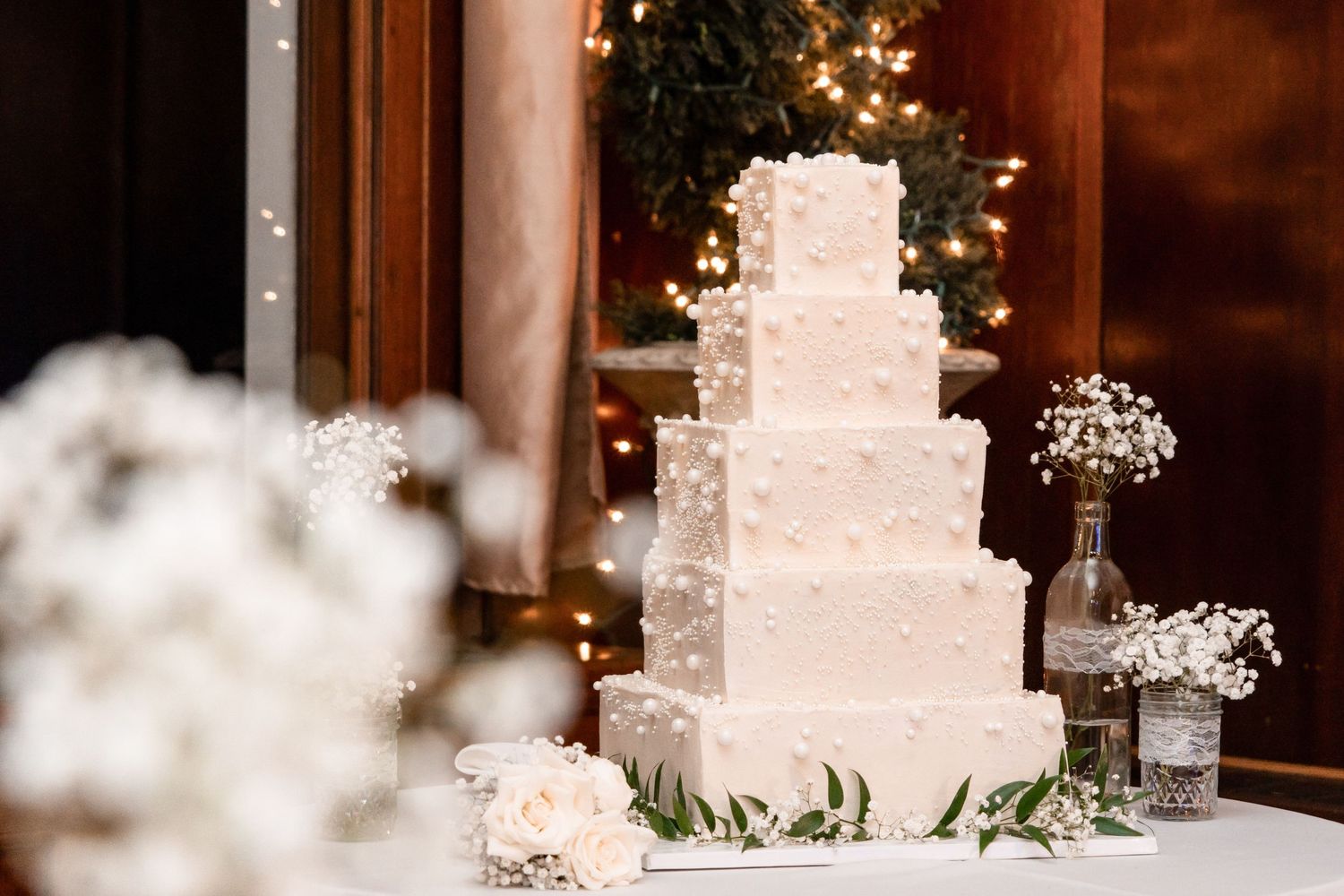 A white elegant wedding cake decorated with delicate floral details and baby's breath sits on a glass display stand.
