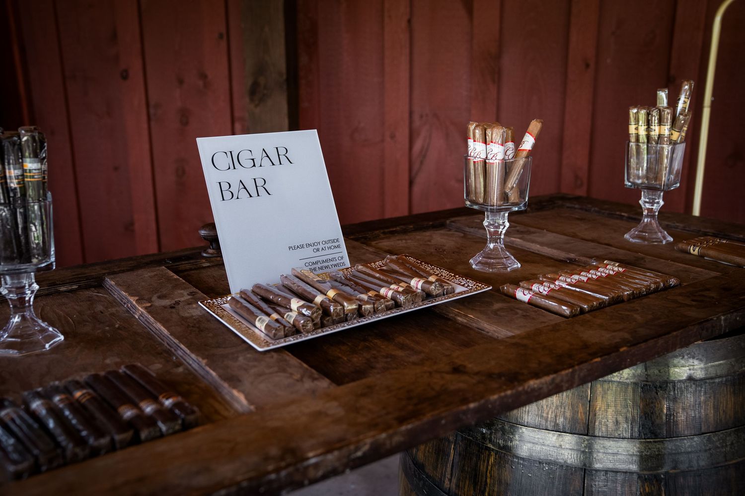 A vintage wooden cigar bar display case with rows of premium cigars and whiskey glasses arranged on a barrel top surface.