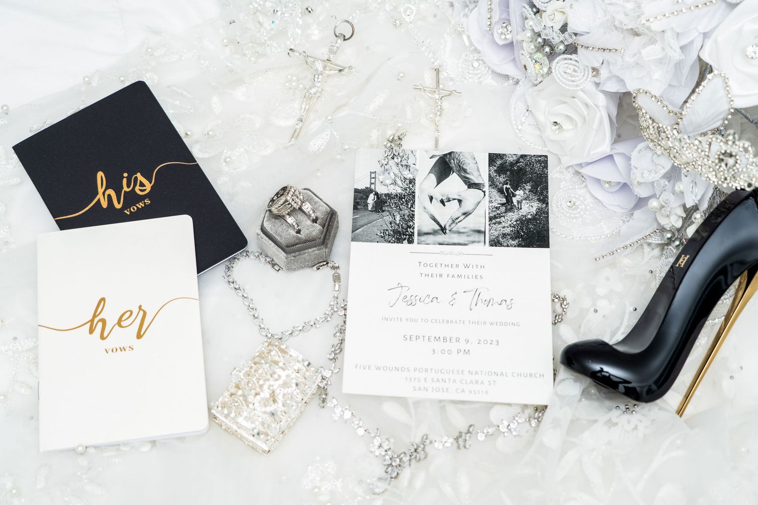 Wedding details flatlay with invitation, black and white notebooks, rhinestone chain, and black stiletto heel on marble surface.