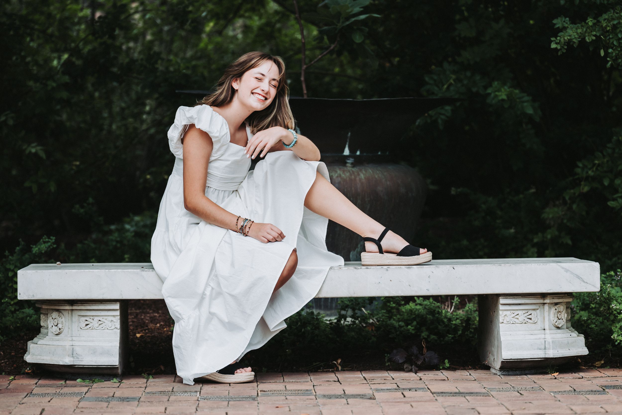 Girl in white dress poses casually on stone bench in garden setting.