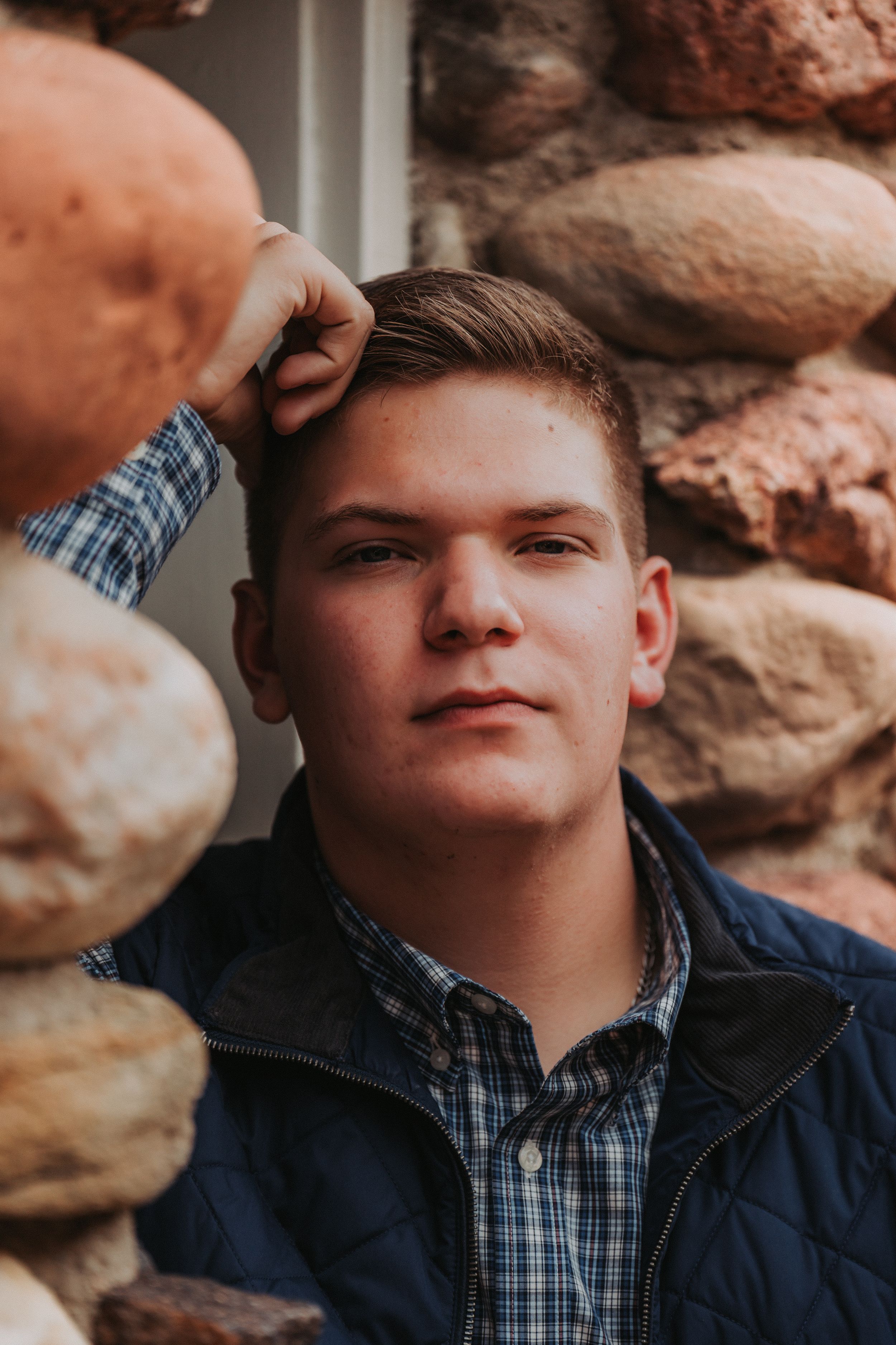 Portrait of a young man in a plaid shirt and navy jacket against a stone wall background.