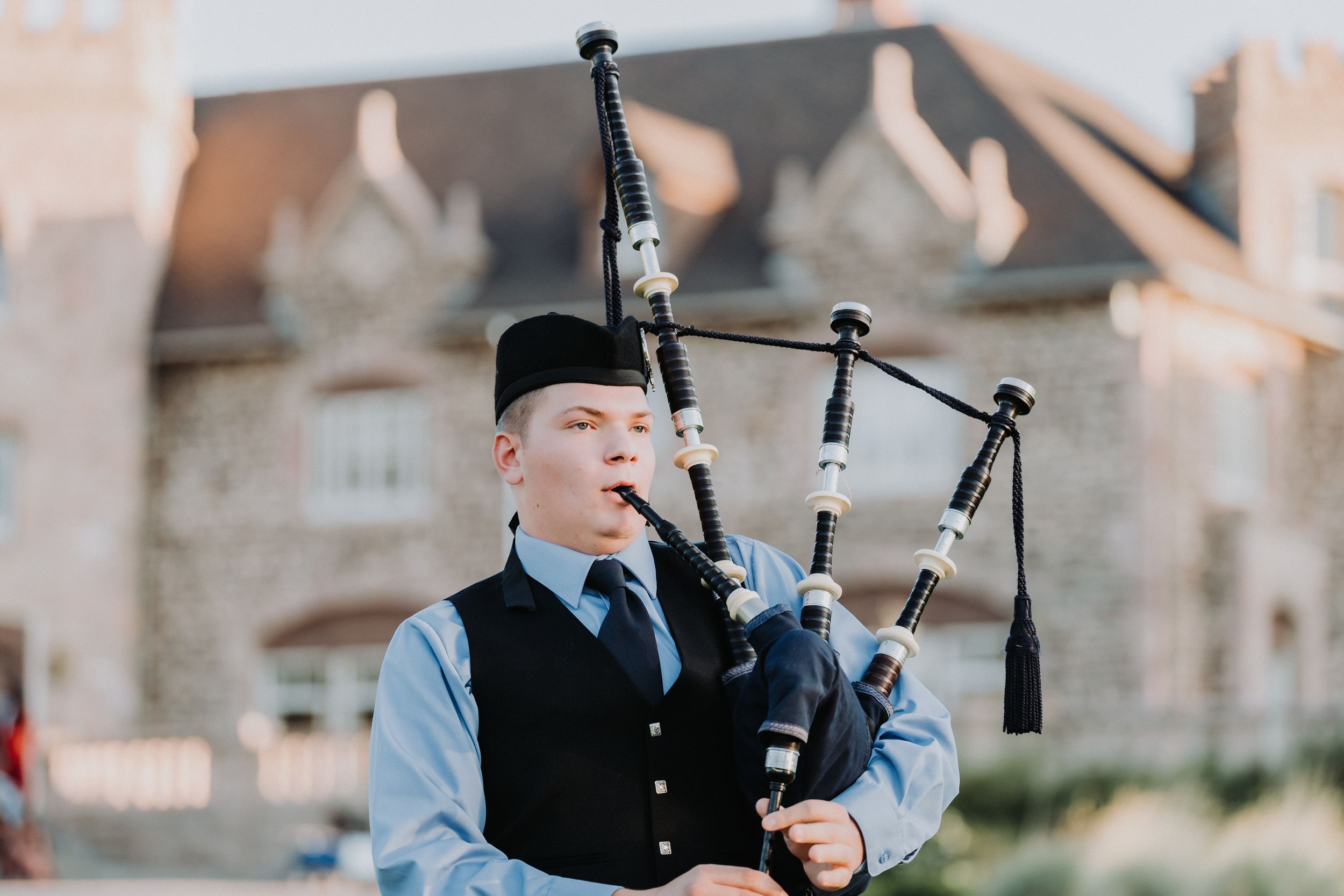 Young bagpiper in traditional dress plays bagpipes outdoors at sunset.