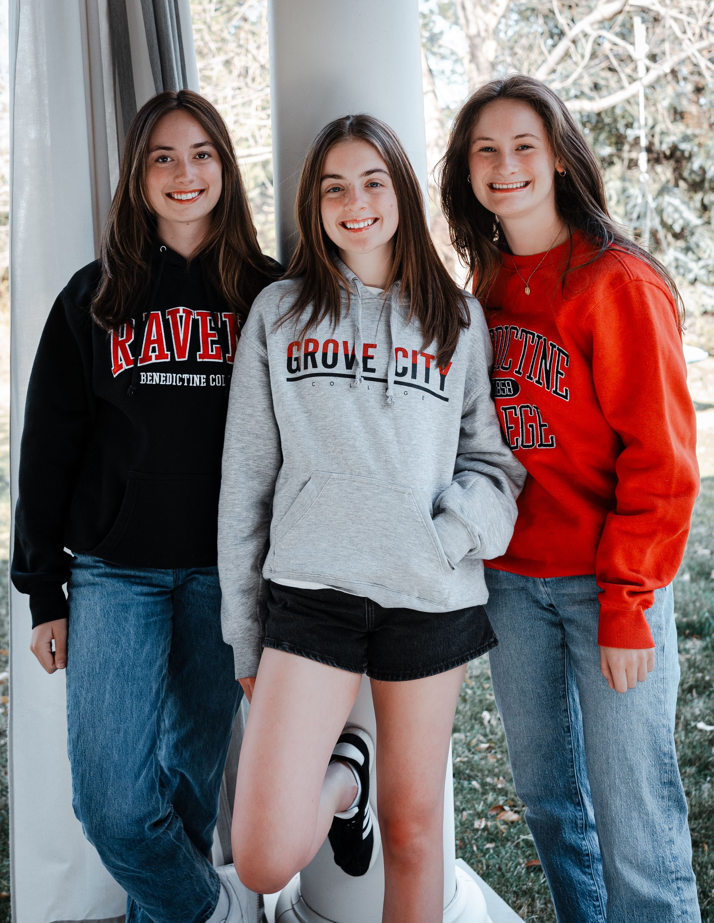 Three friends in college sweatshirts pose together on a porch.