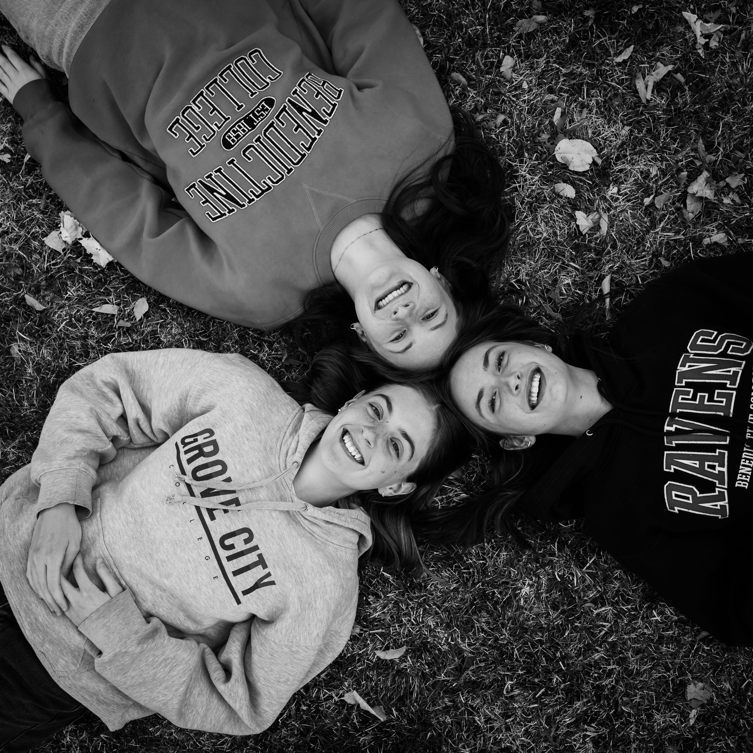 Friends wearing college sweatshirts lie on grass in black and white photo.
