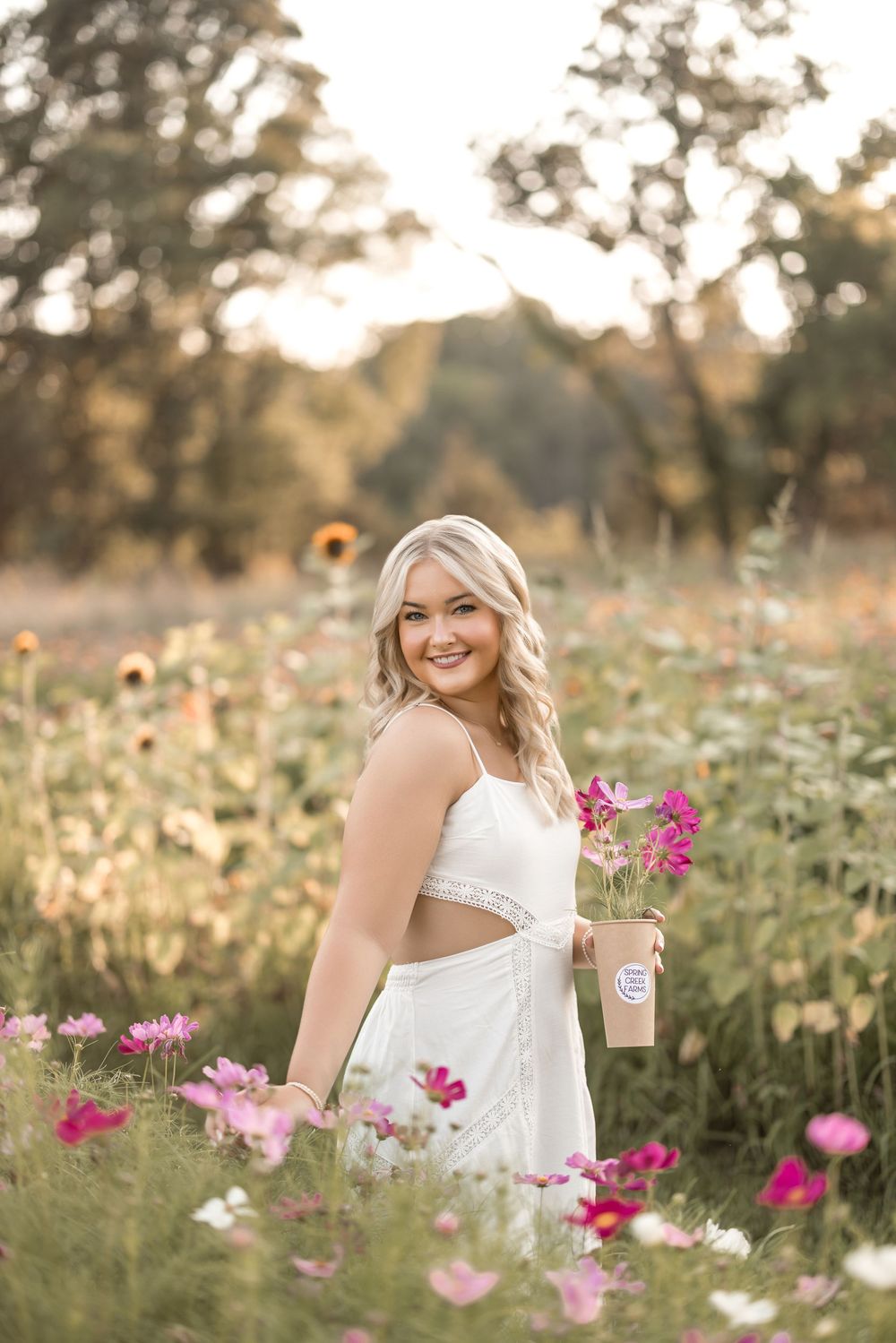 A white dress-clad figure stands in a wildflower field with pink cosmos flowers at golden hour sunset.