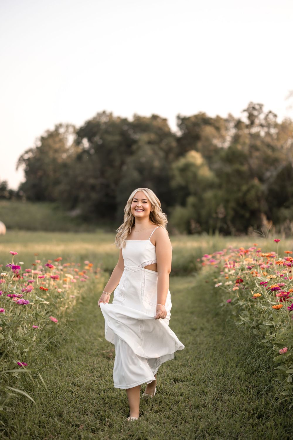 Person in flowing white dress walking along grassy path between colorful wildflower fields at sunset.