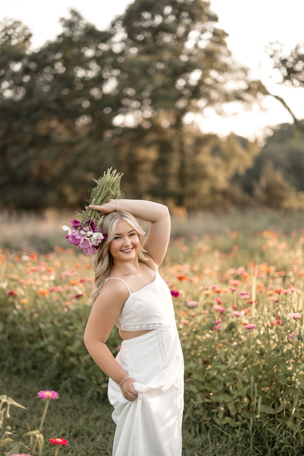 Dreamy summer portrait in a flower field at golden hour featuring white dress and pink wildflower accents.