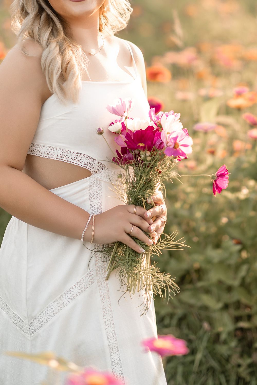 A person in a white lace dress holding a delicate bouquet of pink cosmos flowers in a sunlit garden.