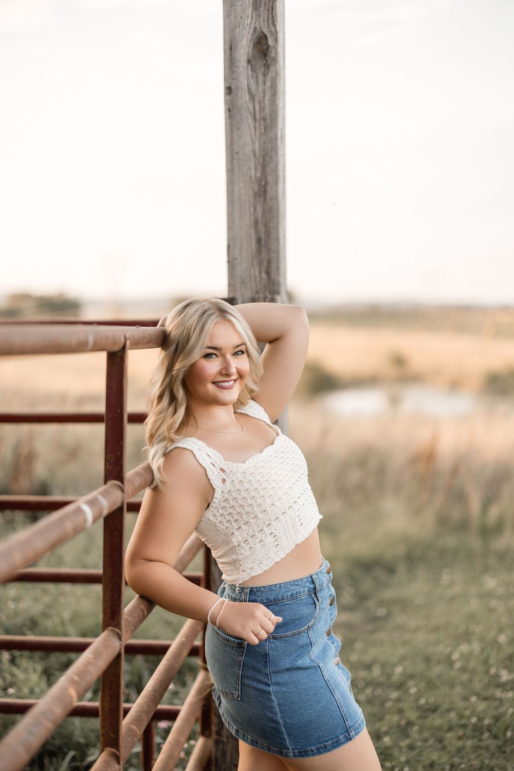 A country photoshoot features a casual summer outfit with a white crochet top and denim skirt by a rustic fence.