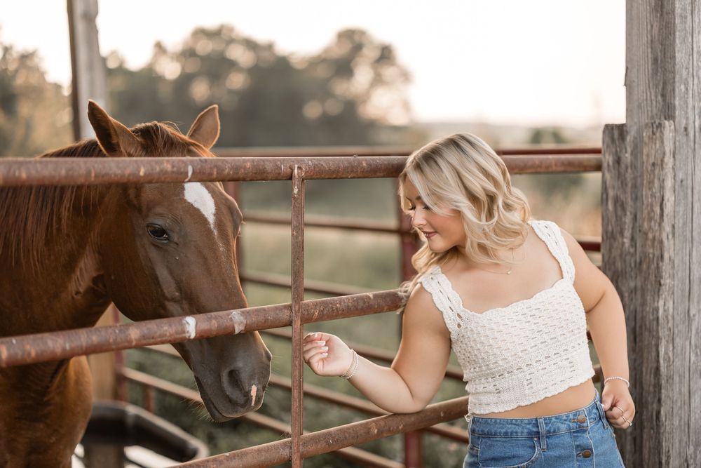 Young equestrian in casual attire bonding with brown horse through rustic metal fence at sunset.