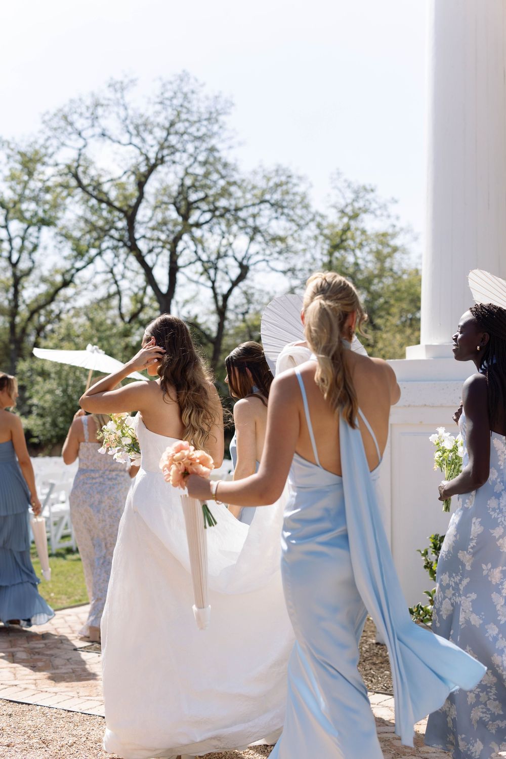 A row of bridesmaids in light blue and white dresses walk away from camera with their hands raised playfully.