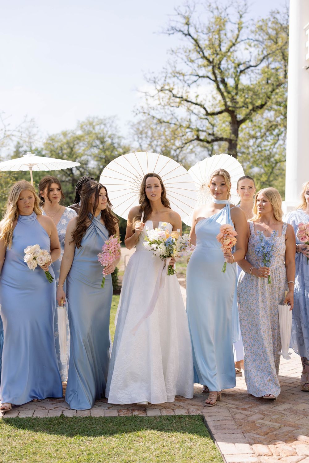 Wedding party dressed in pale blue gowns and white dress pose outdoors with parasol and bouquets on sunny day.